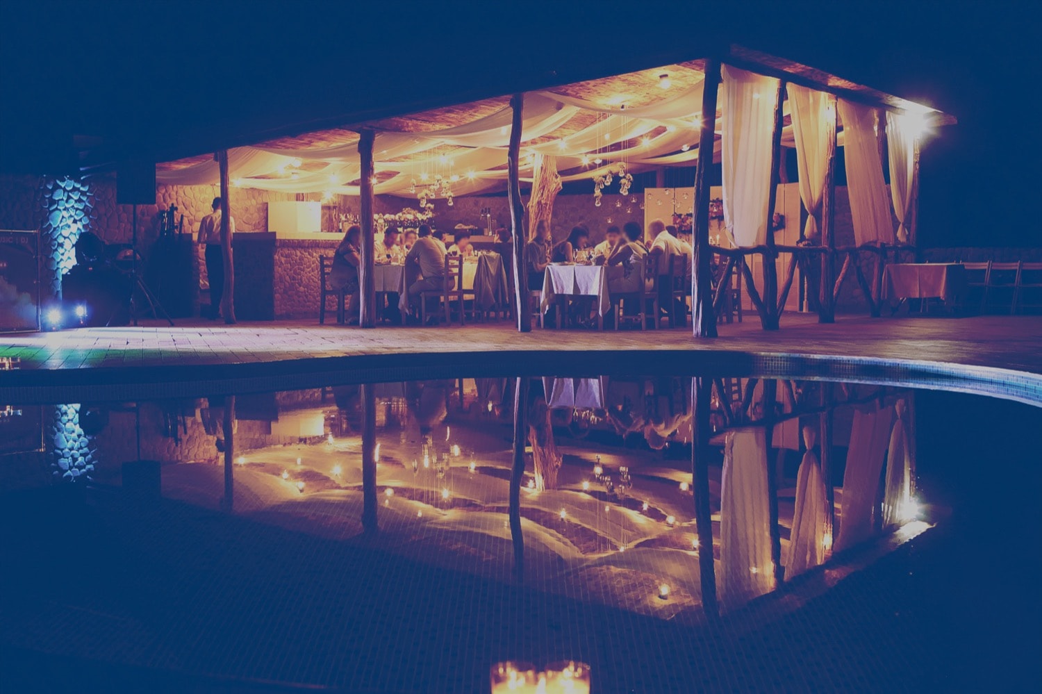 Un grand groupe de personnes mangent le soir dans un restaurant extérieur, à côté d’une piscine.