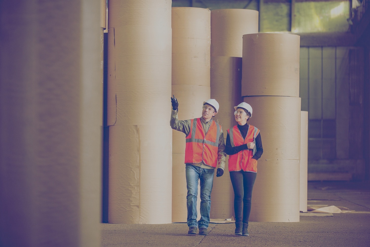 Deux personnes portant un casque traversant une salle industrielle contenant des rouleaux de moquette.