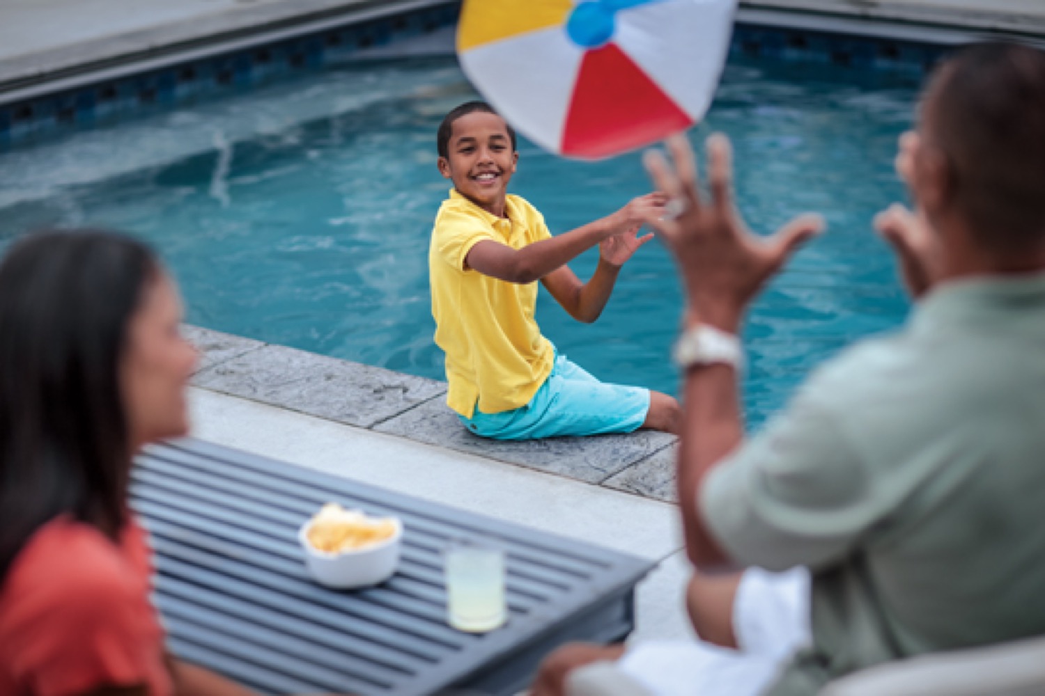 Un garçon souriant assis au bord d'une piscine jette un ballon de plage vers son père.